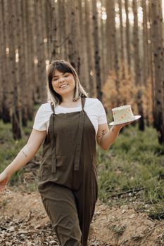 Smiling woman holding a birthday cake in a serene forest setting, capturing a joyful outdoor moment.