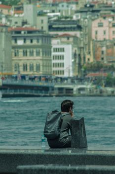 Man with a backpack sits by the water in an urban setting, contemplating life.