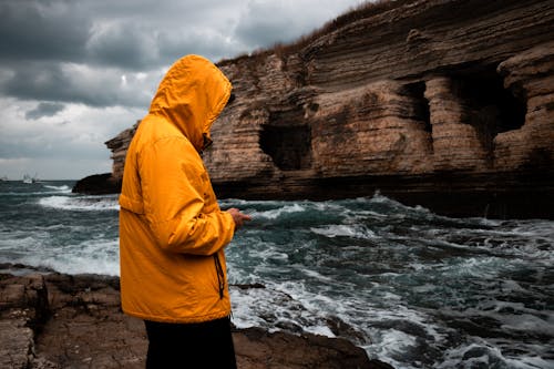 Person Wearing a Yellow Windbreaker Texting During a Storm Standing on Rocky Seashore