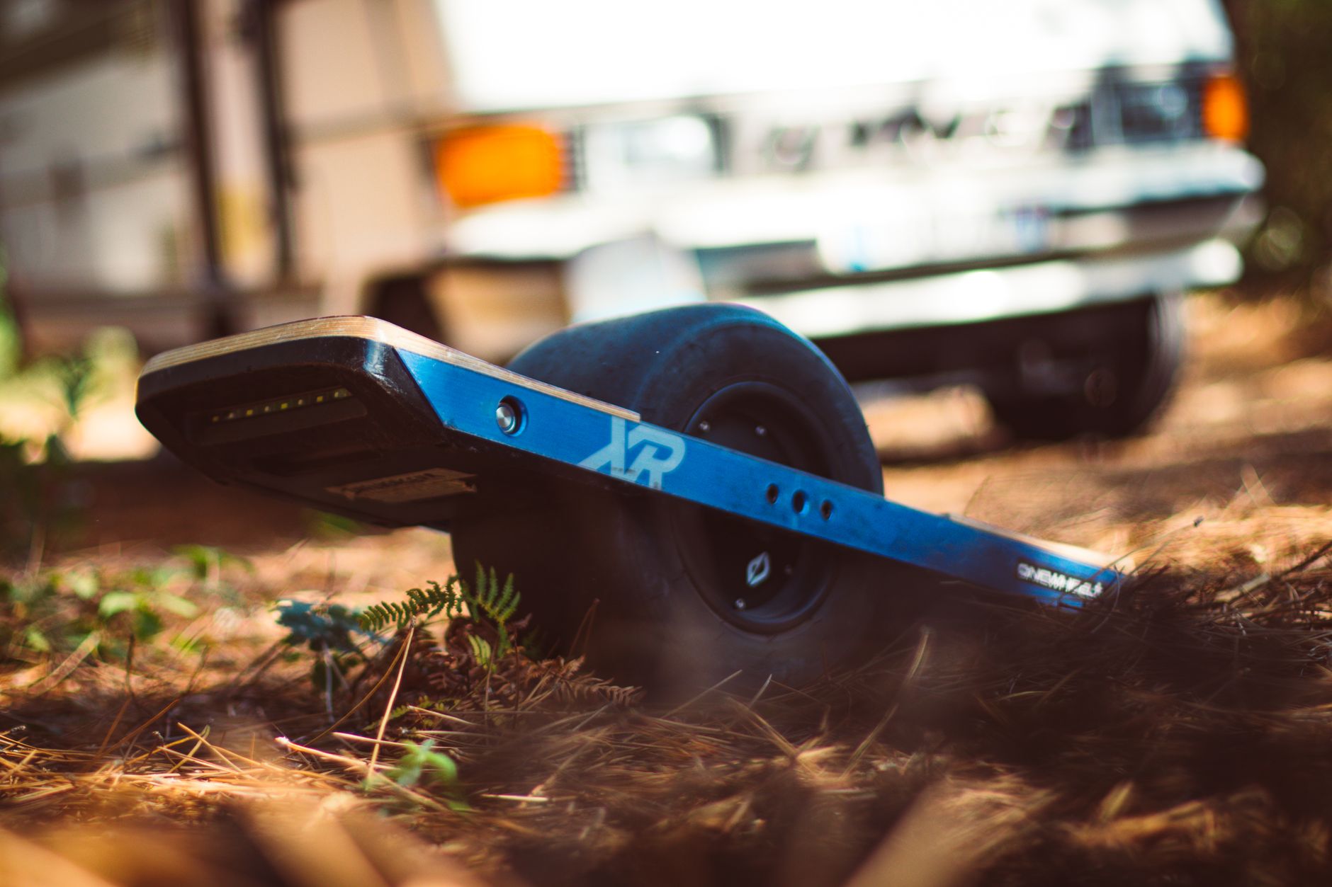 Close-up of a blue Onewheel XR electric skateboard resting on forest ground with a blurred camper in the background.