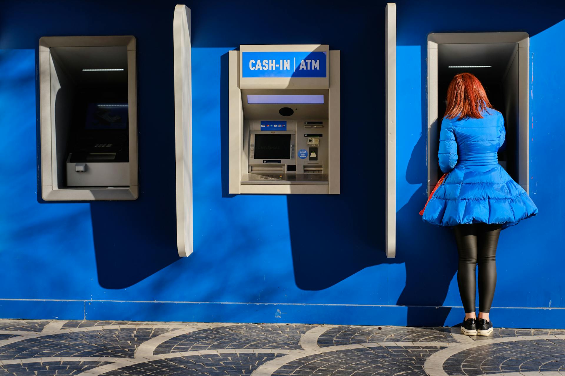 Redheaded woman in blue dress at ATM against vibrant blue wall in Baku, Azerbaijan.