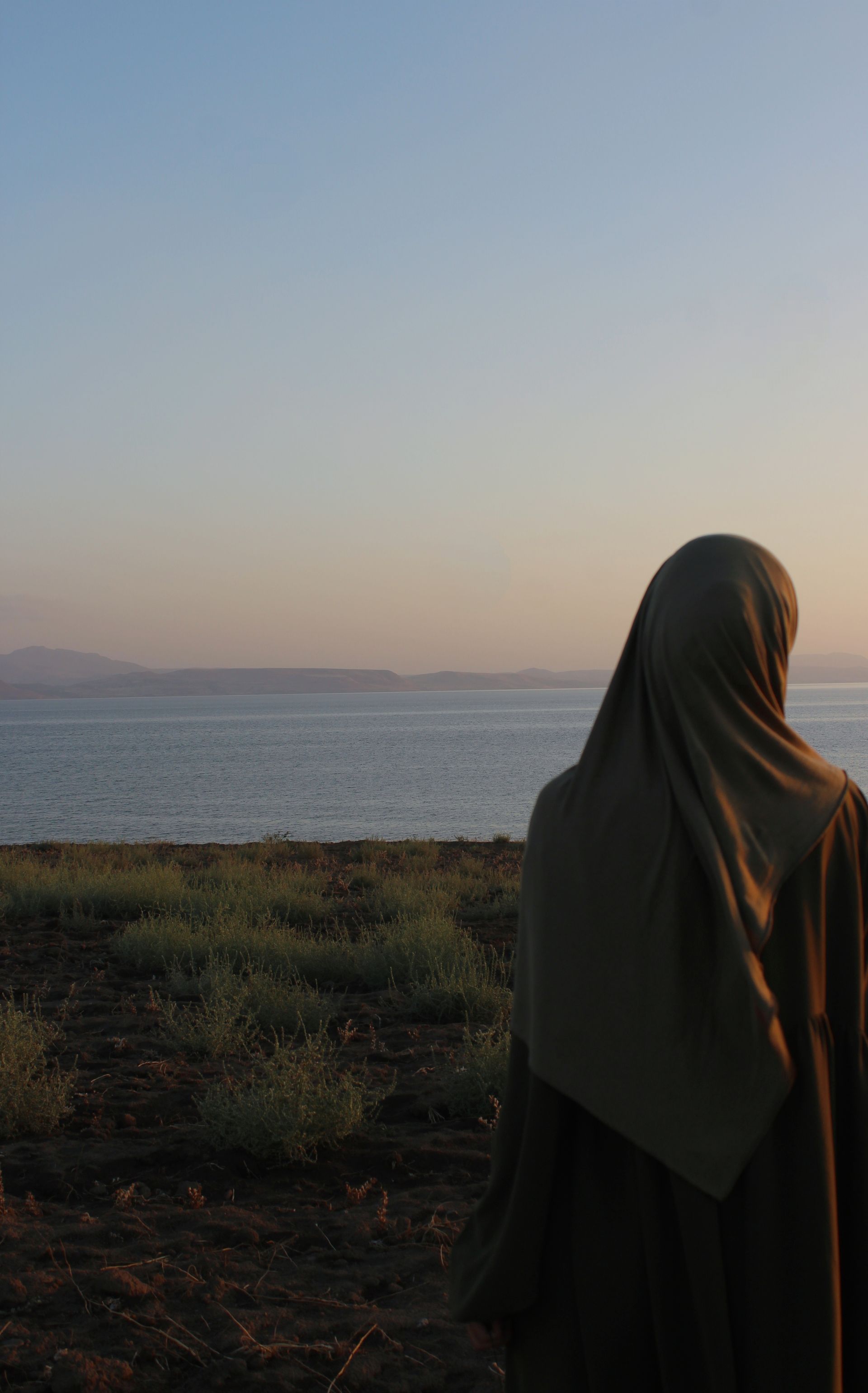 A woman in a hijab gazing at the tranquil sea during sunrise, embracing peace.