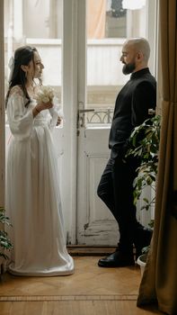 A fashionable wedding couple posing elegantly indoors by a window with plants.