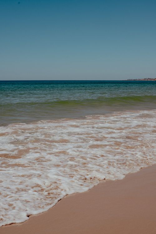 Free Peaceful seashore scene with foamy waves gently lapping against the sandy beach under a clear blue sky. Stock Photo