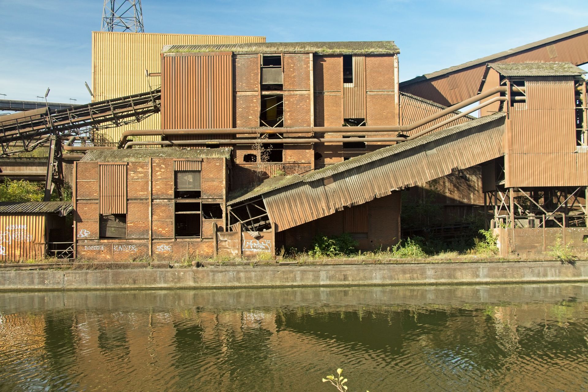 Old industrial factory with rusting metal by a river under clear skies.