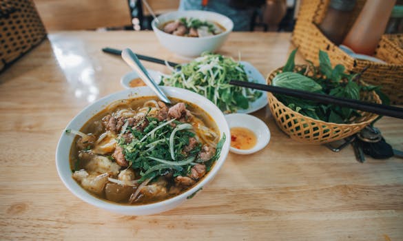 A vibrant bowl of Vietnamese pho topped with fresh greens, surrounded by herbs on a wooden table.