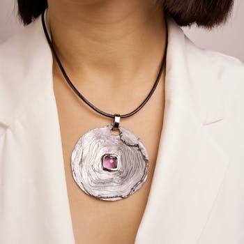 Close-up of an elegant silver pendant with a purple gemstone worn by a woman in a white blazer.