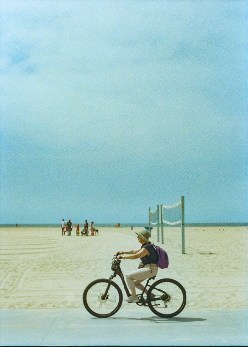 Free Woman riding bicycle on beachside path with volleyball net and people in background. Stock Photo