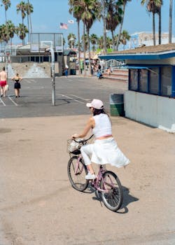 A woman rides a bicycle along a palm tree-lined path near a beachside basketball court.