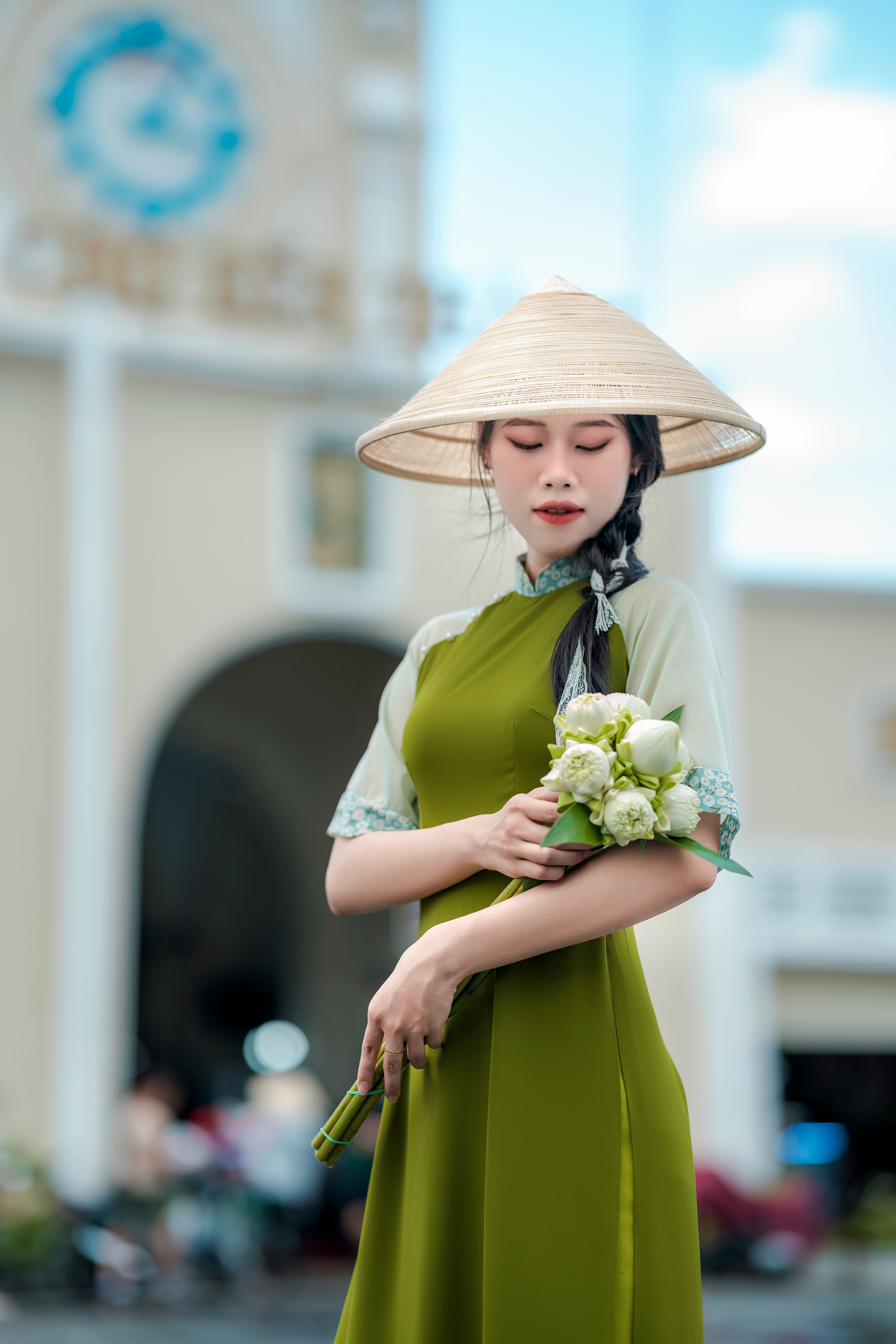 A Vietnamese woman in a traditional green dress with conical hat holding flowers outdoors.