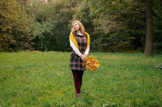 A woman enjoys an autumn day in a forest, holding golden leaves.
