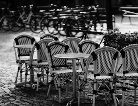 Charming black and white photo of outdoor cafe seating in an urban square, featuring woven chairs and bicycles.