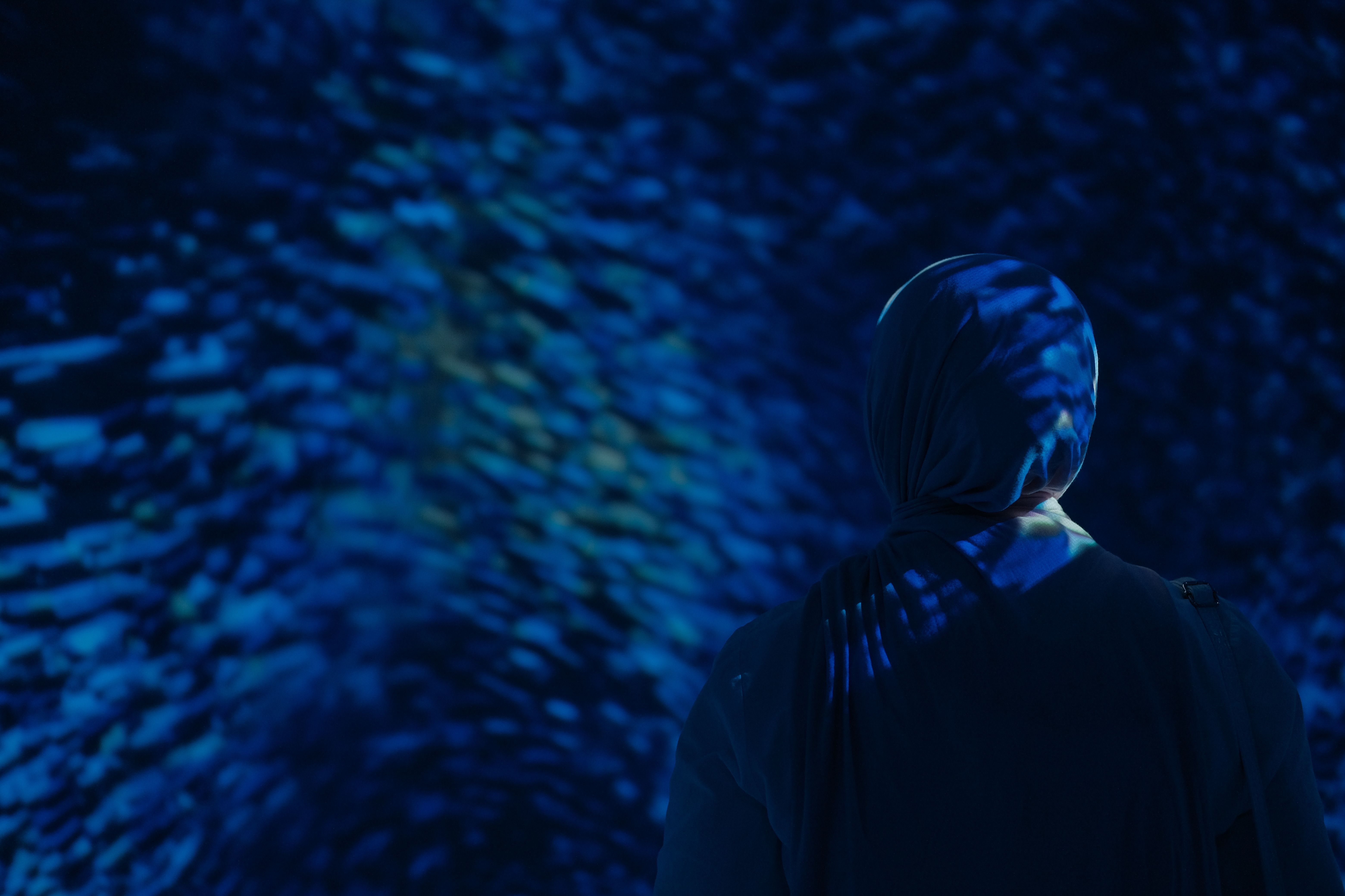A Muslim woman in a hijab standing in a dark room with blue aquarium lighting.