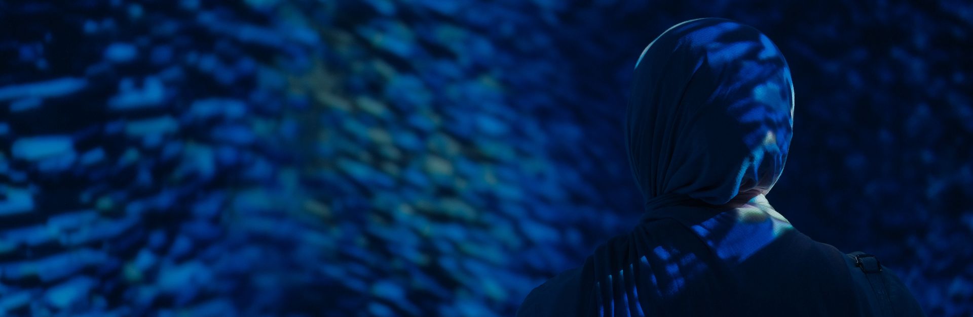 A Muslim woman in a hijab standing in a dark room with blue aquarium lighting.