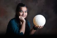 A young woman poses with a glowing moon model in a studio setting, exuding warmth and creativity.