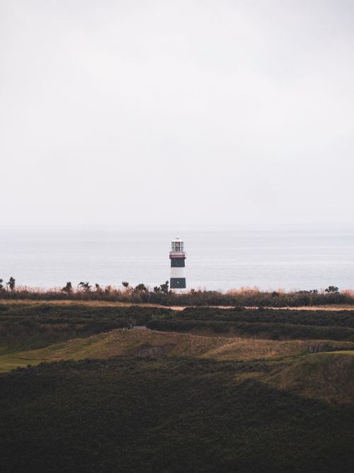 Free A picturesque lighthouse stands tall against a calm sea and overcast sky, surrounded by lush greenery. Stock Photo