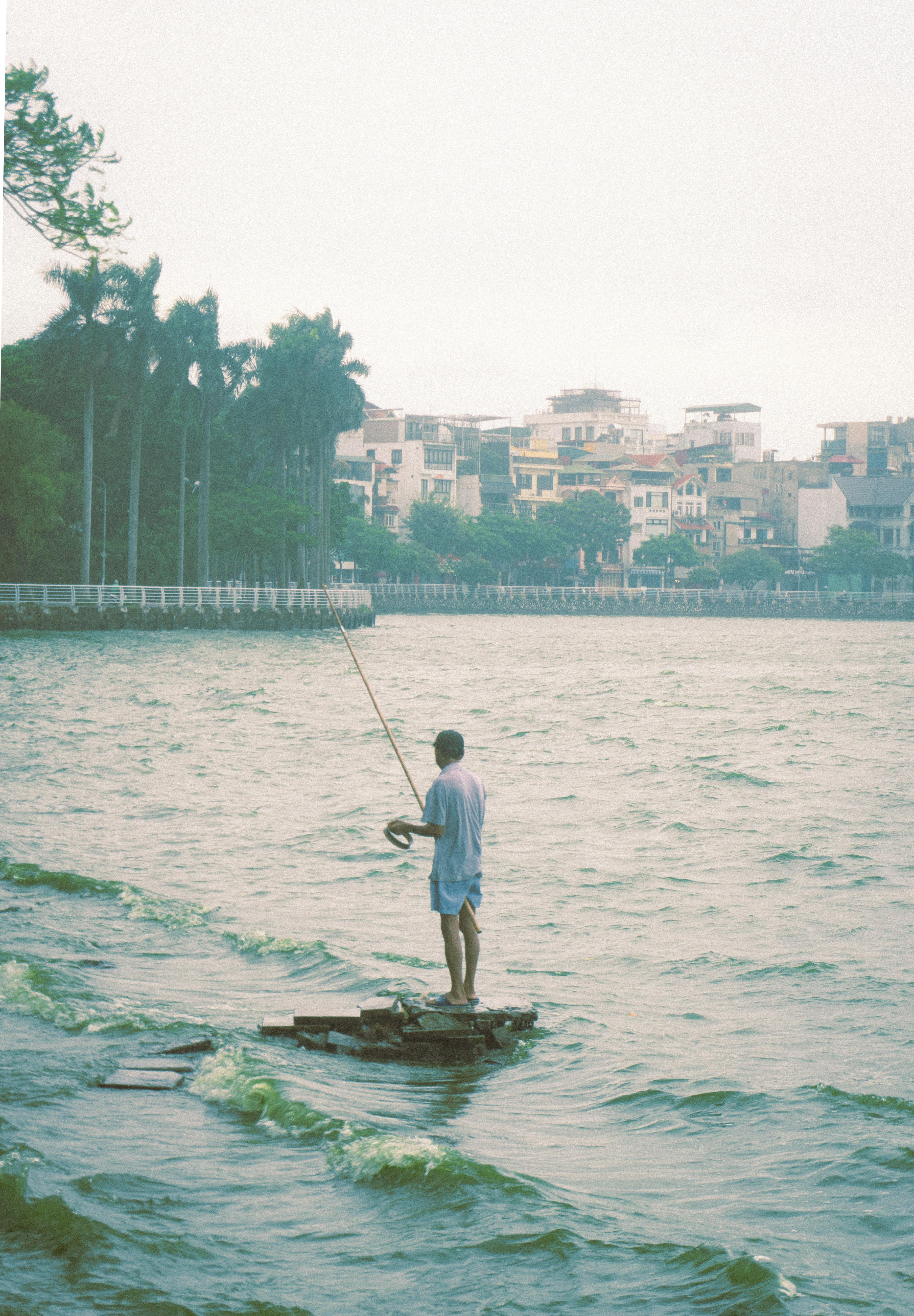 A lone fisherman stands in a lake in Hanoi, Vietnam, casting his line amid urban scenery.