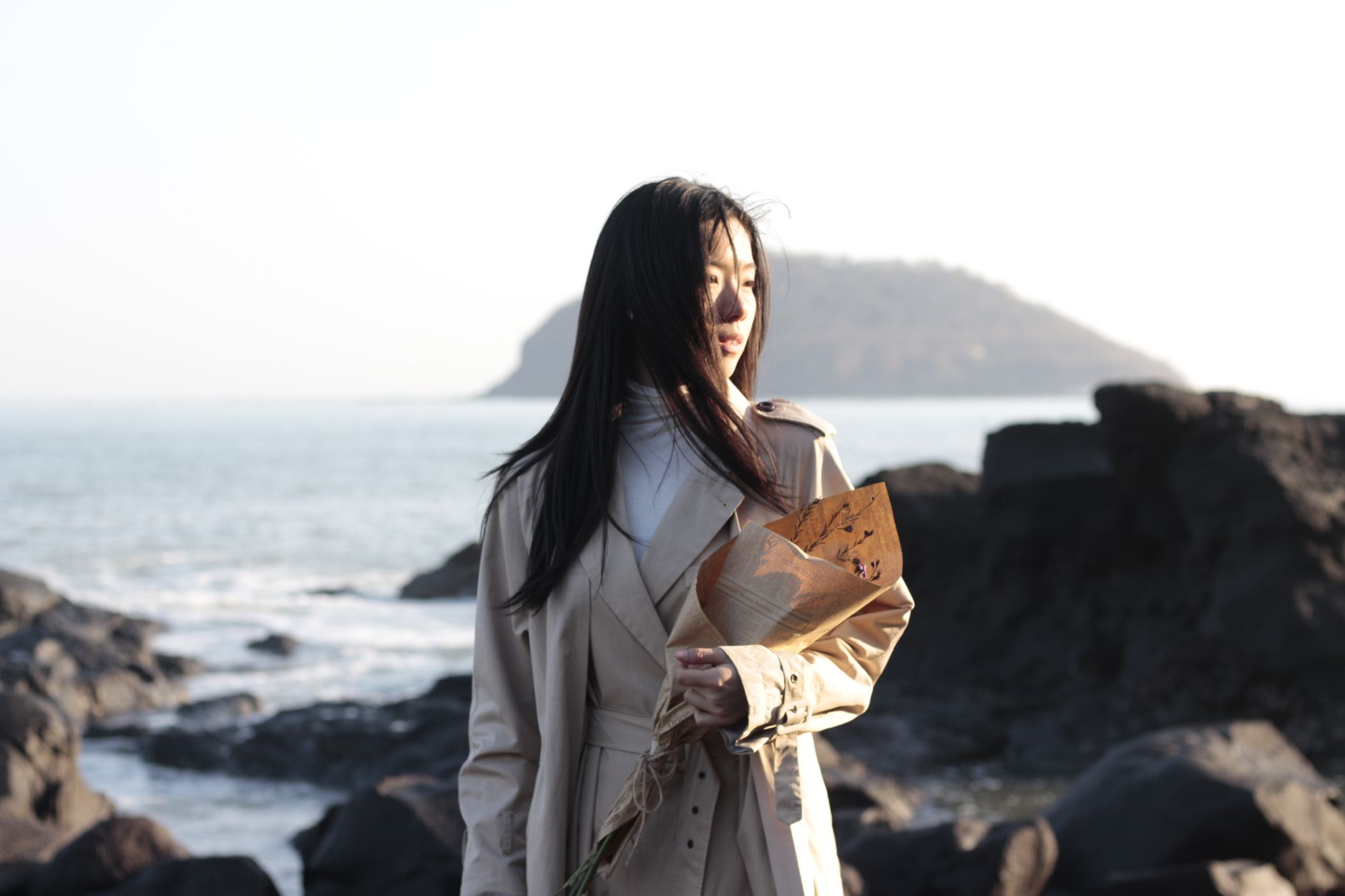 Asian woman in a trench coat holding flowers on a rocky seashore with a distant island view.