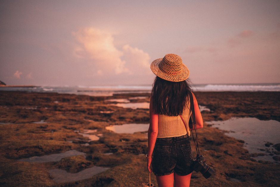 A woman in a straw hat gazes at the ocean during a beautiful sunset on a rocky beach.