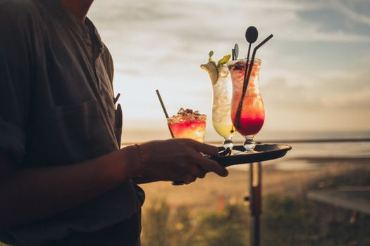 A server carries colorful cocktails at a beachside sunset setting, perfect for a tropical getaway vibe.