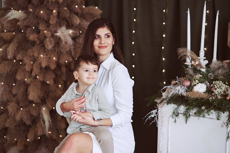 Smiling Woman With Her Little Son On Her Lap Sitting Next To The Christmas Tree