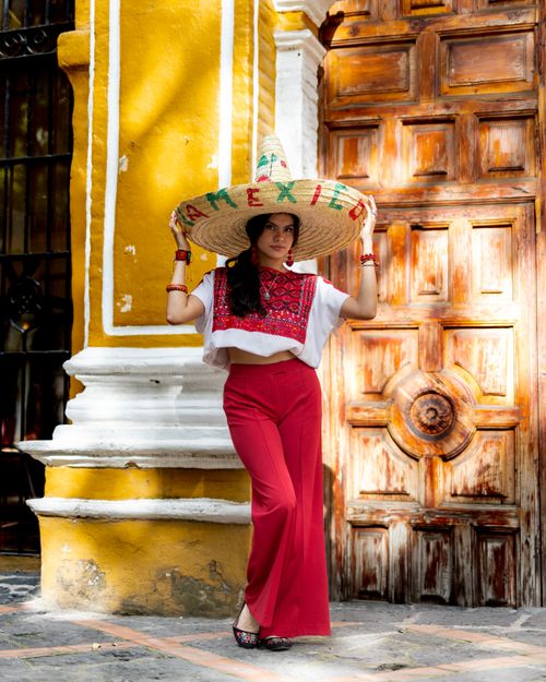 Free Woman in traditional Mexican clothing and sombrero, standing by an ornate wooden door with vibrant yellow wall. Stock Photo