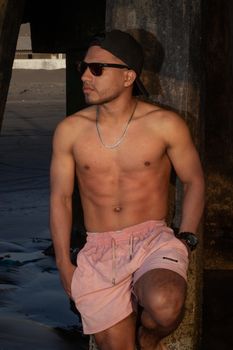A fit and muscular man wearing sunglasses and shorts stands under a pier at sunset, showcasing strength and style.