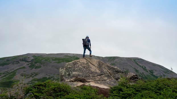 A lone hiker stands atop a rock with a scenic mountain backdrop.