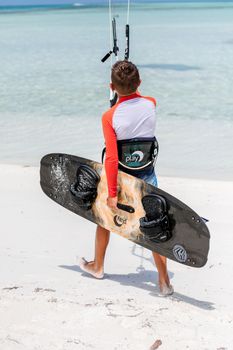 Teen boy holding a kiteboard on a sunny beach, ready for kitesurfing adventure.