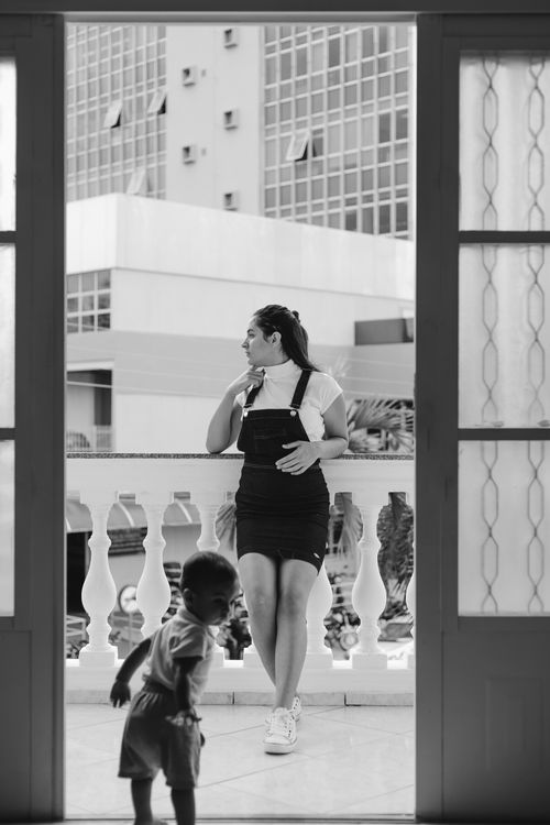 Free Woman posing on a balcony as a child plays nearby in a cityscape. Stock Photo
