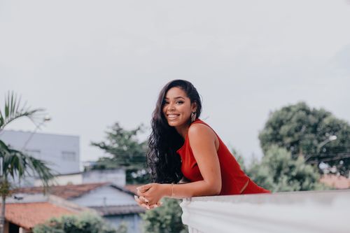 Free Smiling woman in a red dress enjoying a sunny day outdoors, leaning on a white railing. Stock Photo
