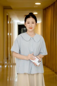 Smiling woman holding a smartphone while standing in a well-lit hallway.