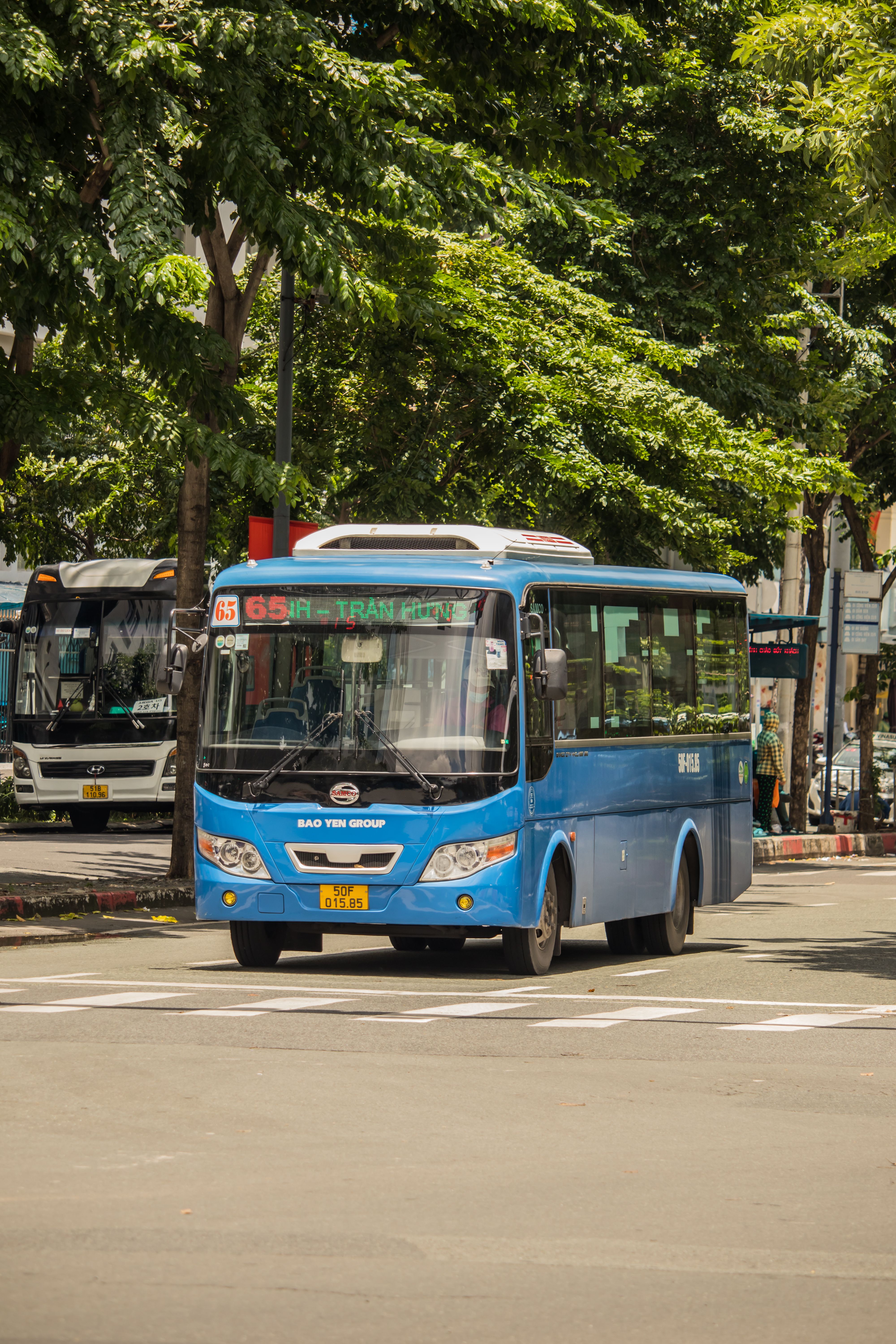 Blue public bus on city street in Ho Chi Minh, Vietnam surrounded by trees and urban scenery.