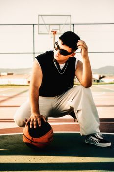 A man squatting with a basketball on an outdoor urban court wearing sunglasses and casual attire.