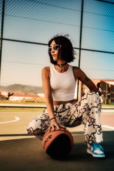 Young woman in trendy outfit posing with basketball at outdoor court.