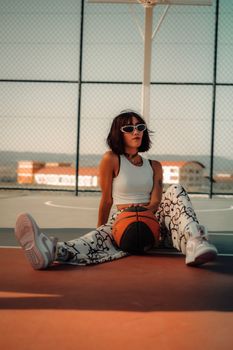 Trendy woman with sunglasses posing on outdoor basketball court with a ball.