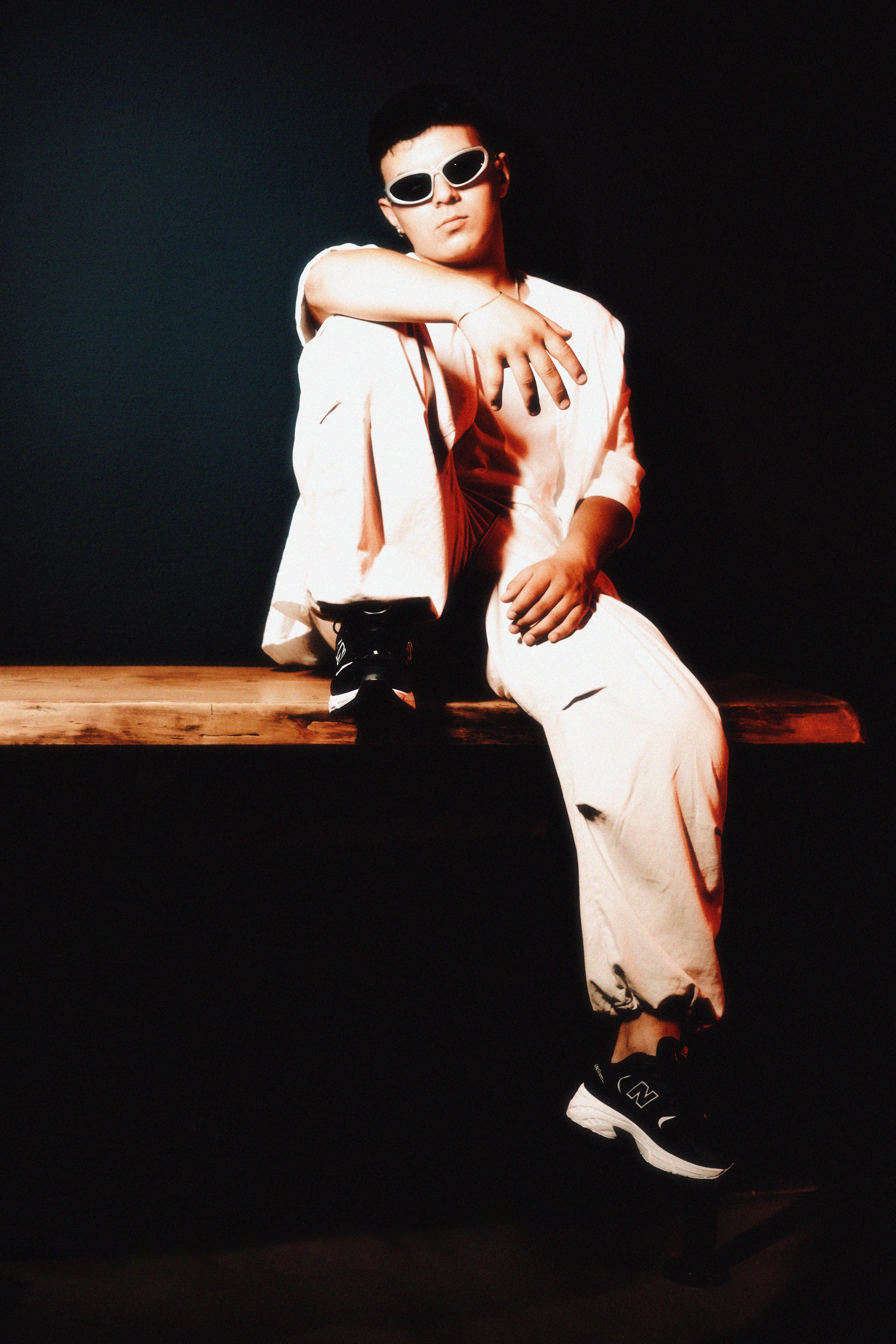 A young man poses stylishly in sunglasses and white outfit, exuding a cool fashion vibe.