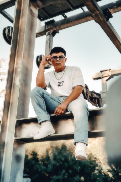 Free A stylish young man poses outdoors in sunglasses, sitting confidently on a steel beam. Stock Photo