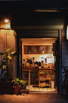 Warmly lit night view of a cozy urban cafe entrance with potted plants and menu chalkboards.