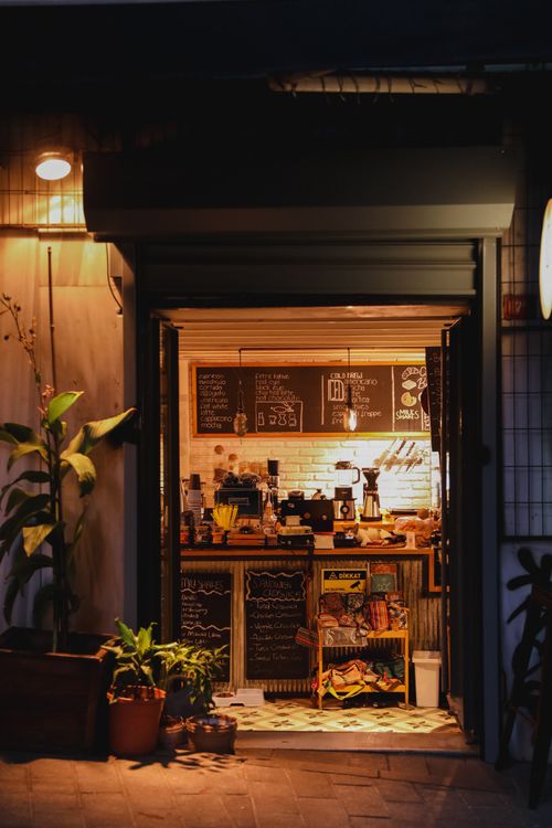 Free Warmly lit night view of a cozy urban cafe entrance with potted plants and menu chalkboards. Stock Photo