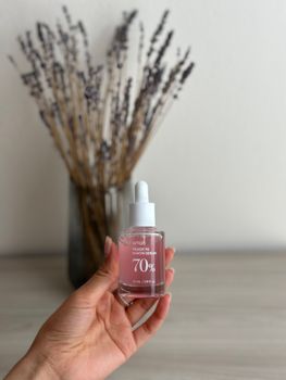 A close-up of a hand holding a serum bottle against a lavender background on a wooden table.