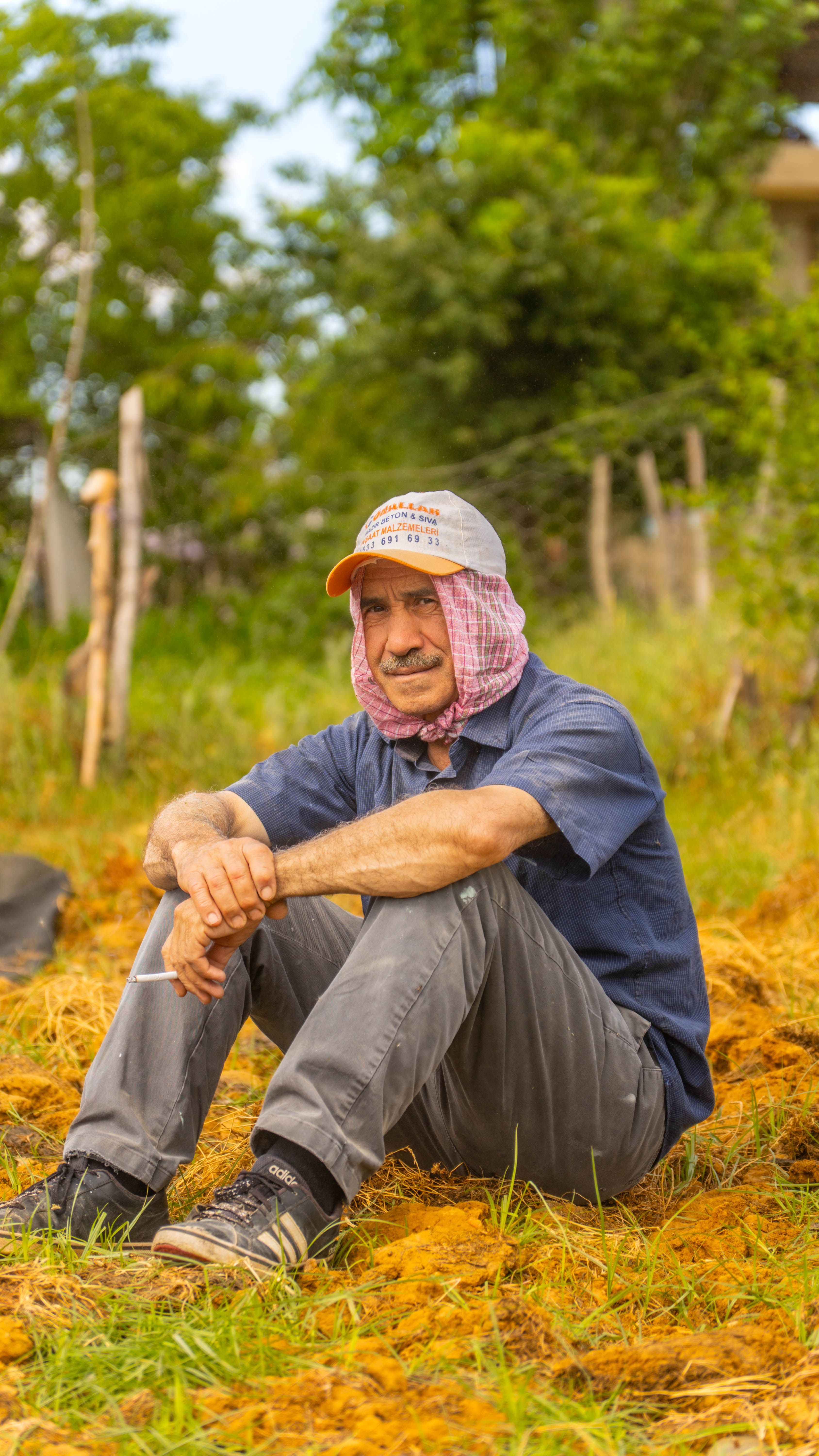 Portrait of a man wearing a baseball cap and scarf, sitting outdoors in a rural area.