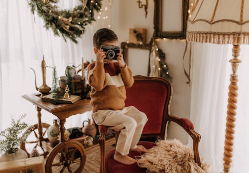Boy in Sweater Posing on Armchair with Camera in Hands
