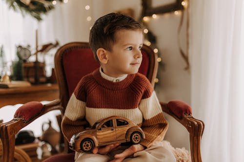 Little Boy in Sweater Sitting on Armchair with Wooden Car in Hands