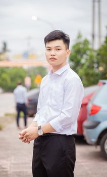 A young Asian man in a white shirt standing confidently outdoors in a parking area on a bright day.