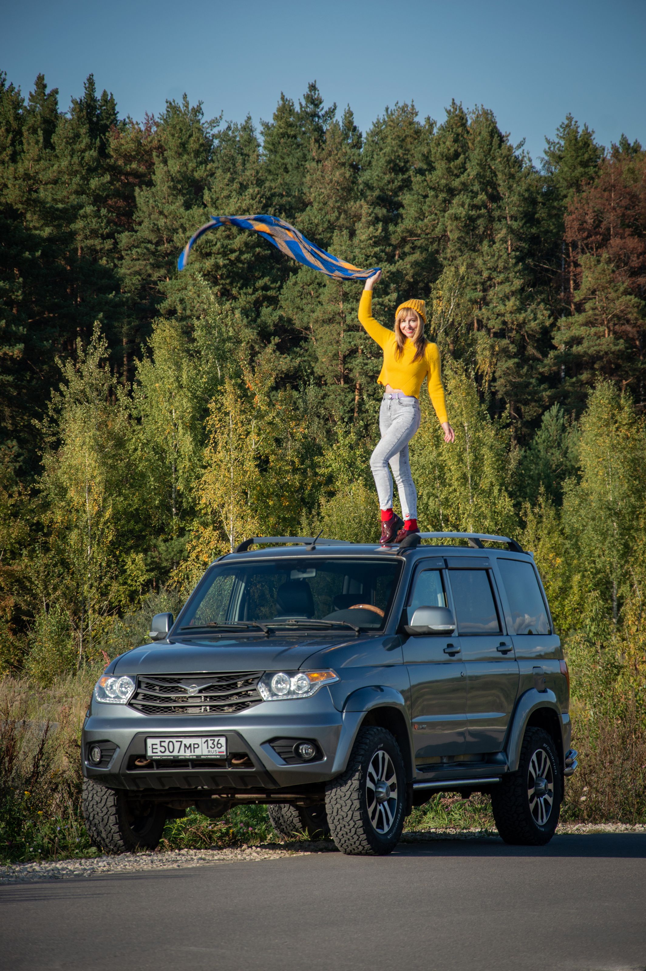 Woman joyfully standing on SUV with colorful scarf in forest setting.