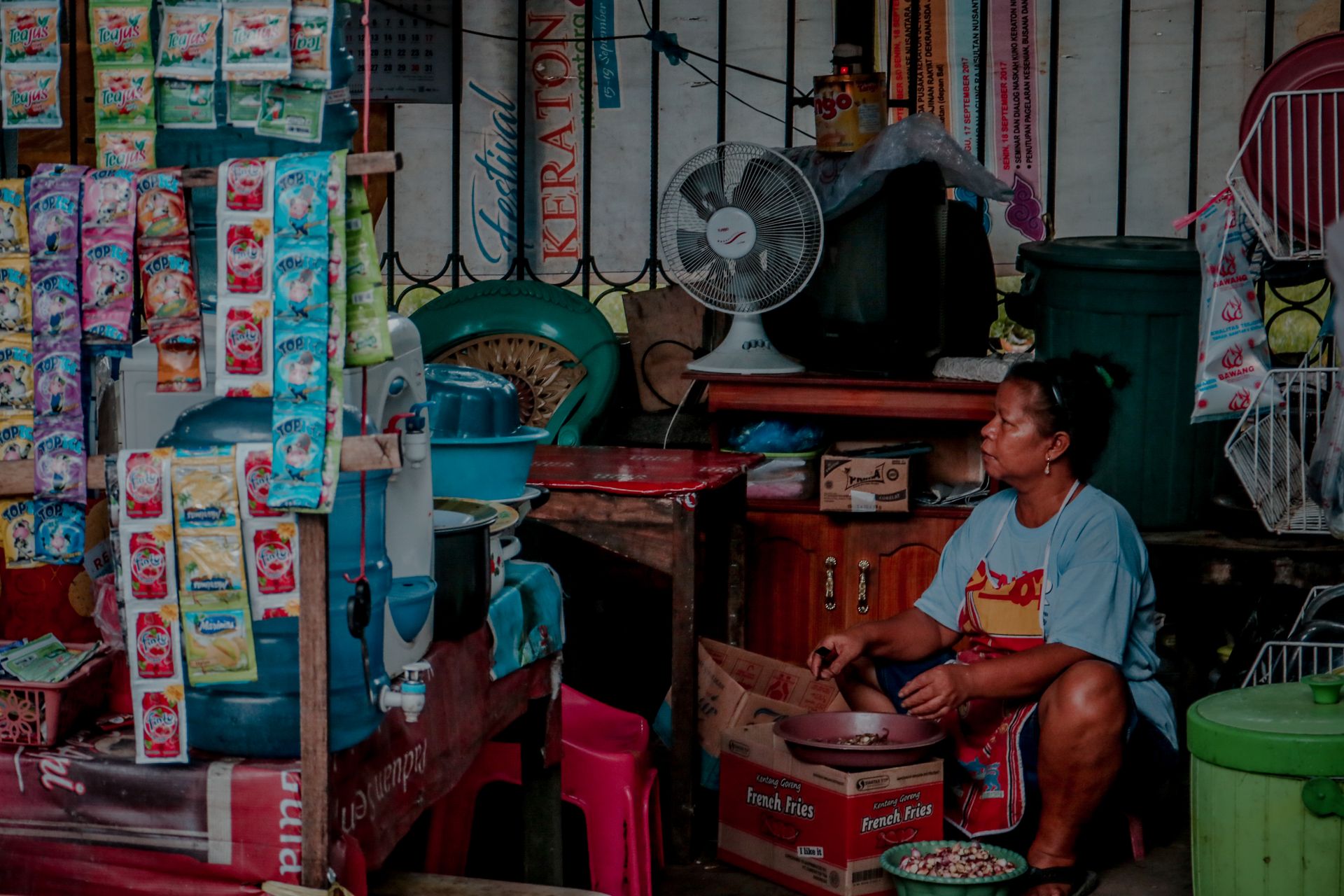 A woman sits in a small retail shop filled with hanging products and food items.