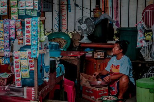 Free A woman sits in a small retail shop filled with hanging products and food items. Stock Photo