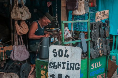 Free A local vendor repairing shoes at a bustling marketplace stall with visible signs and merchandise. Stock Photo