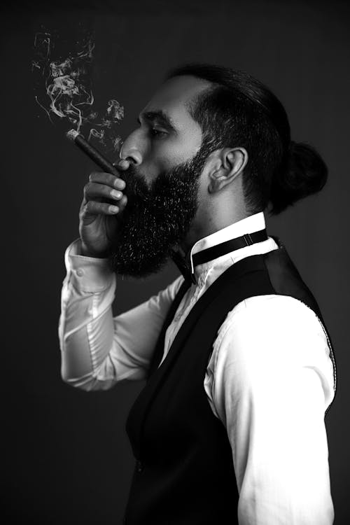 Black and White Photo of a Brunette Bearded Man in Vest and Bow-Tie Smoking a Cigar
